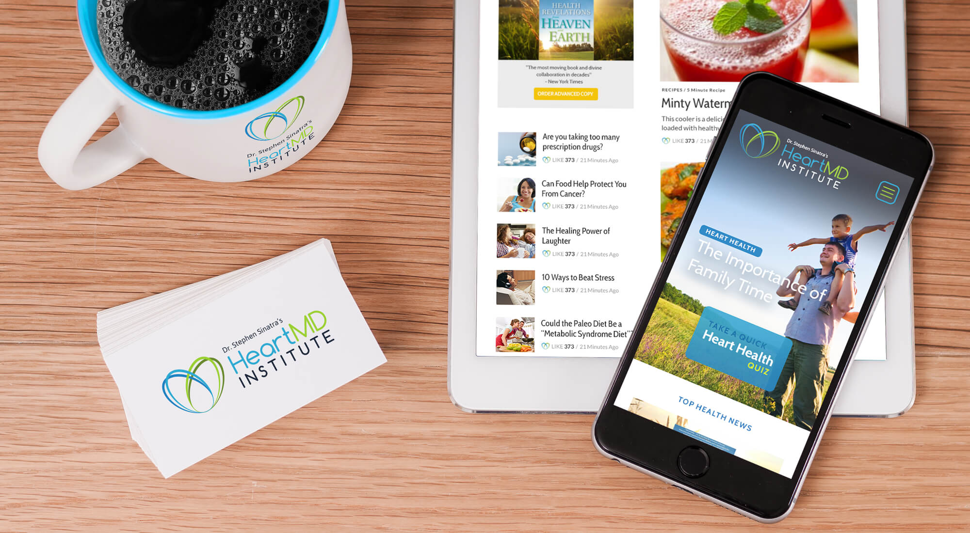 A coffee mug, business cards, tablet, and smartphone with Heart MD Institute branding are arranged on a wooden desk; tablet and phone display the institute’s website.