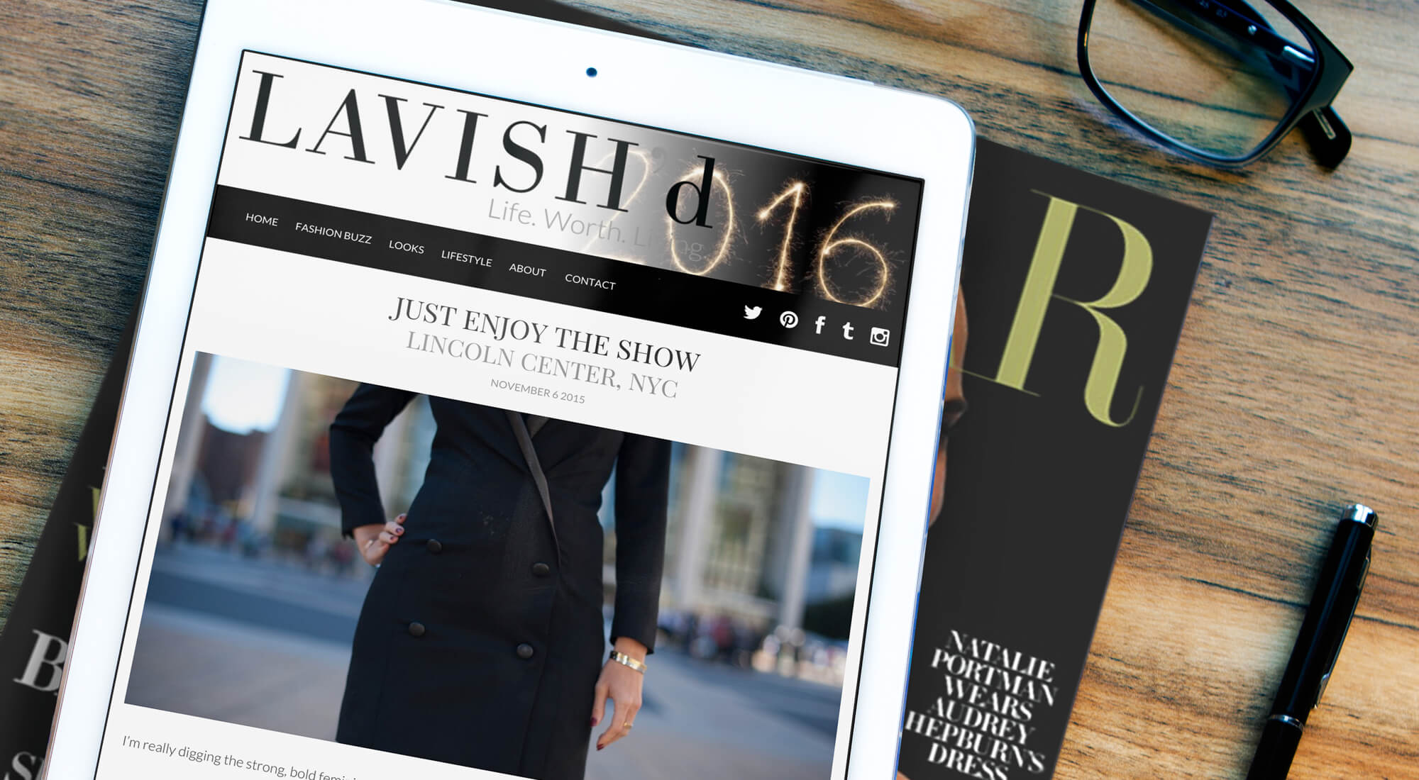 A tablet displays a fashion blog article titled "Just Enjoy the Show, Lincoln Center, NYC" beside a magazine, eyeglasses, and a pen on a wooden surface.