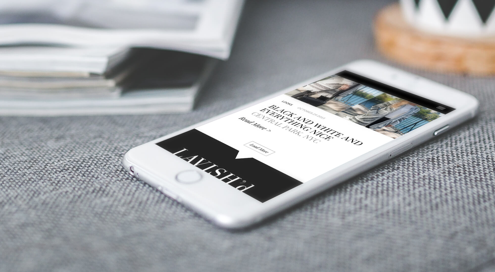 A smartphone rests on a gray fabric surface displaying a website with the title "BLACK AND WHITE AND EVERYTHING NICE." A blurred stack of magazines is in the background.