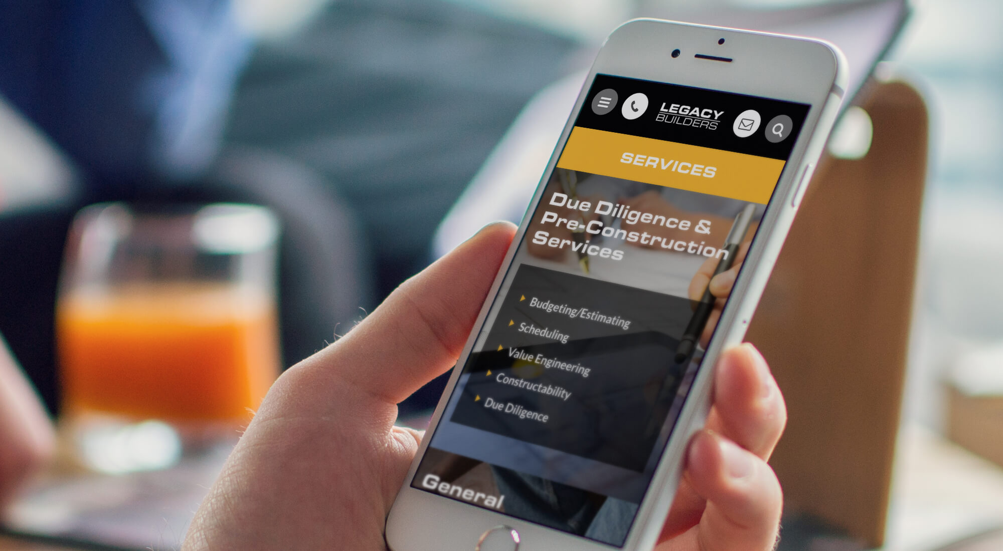 A person holds a smartphone displaying the Legacy Builders website, showing a menu with "Due Diligence & Pre-Construction Services." A glass of orange juice is in the background.