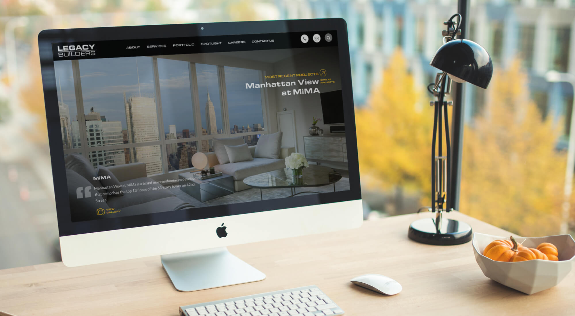 An iMac on a desk displays a website for Legacy Builders, featuring a luxury Manhattan apartment. A lamp, cup, and a bowl of oranges are also on the desk, with city buildings visible outside.