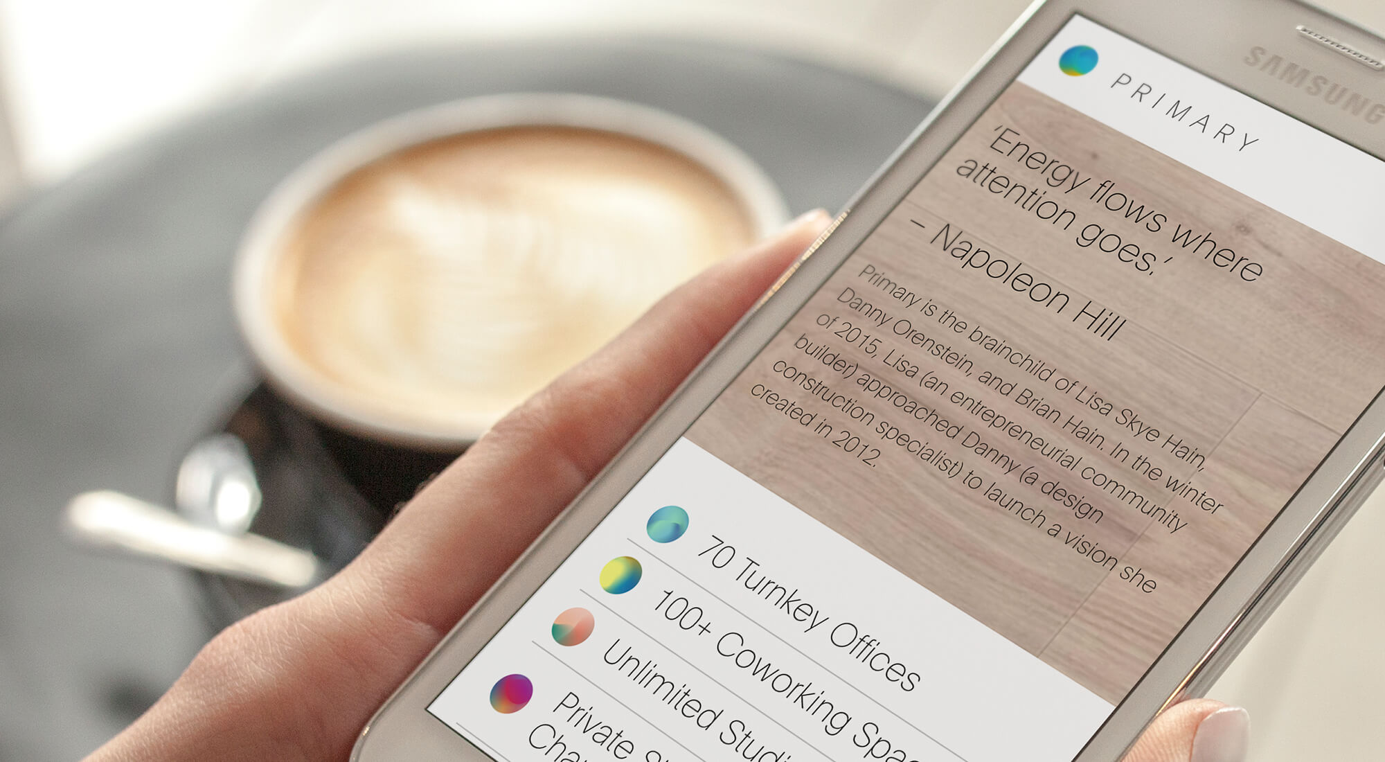 A hand holds a Samsung smartphone displaying a coworking space website, with a coffee cup on a saucer in the blurred background.