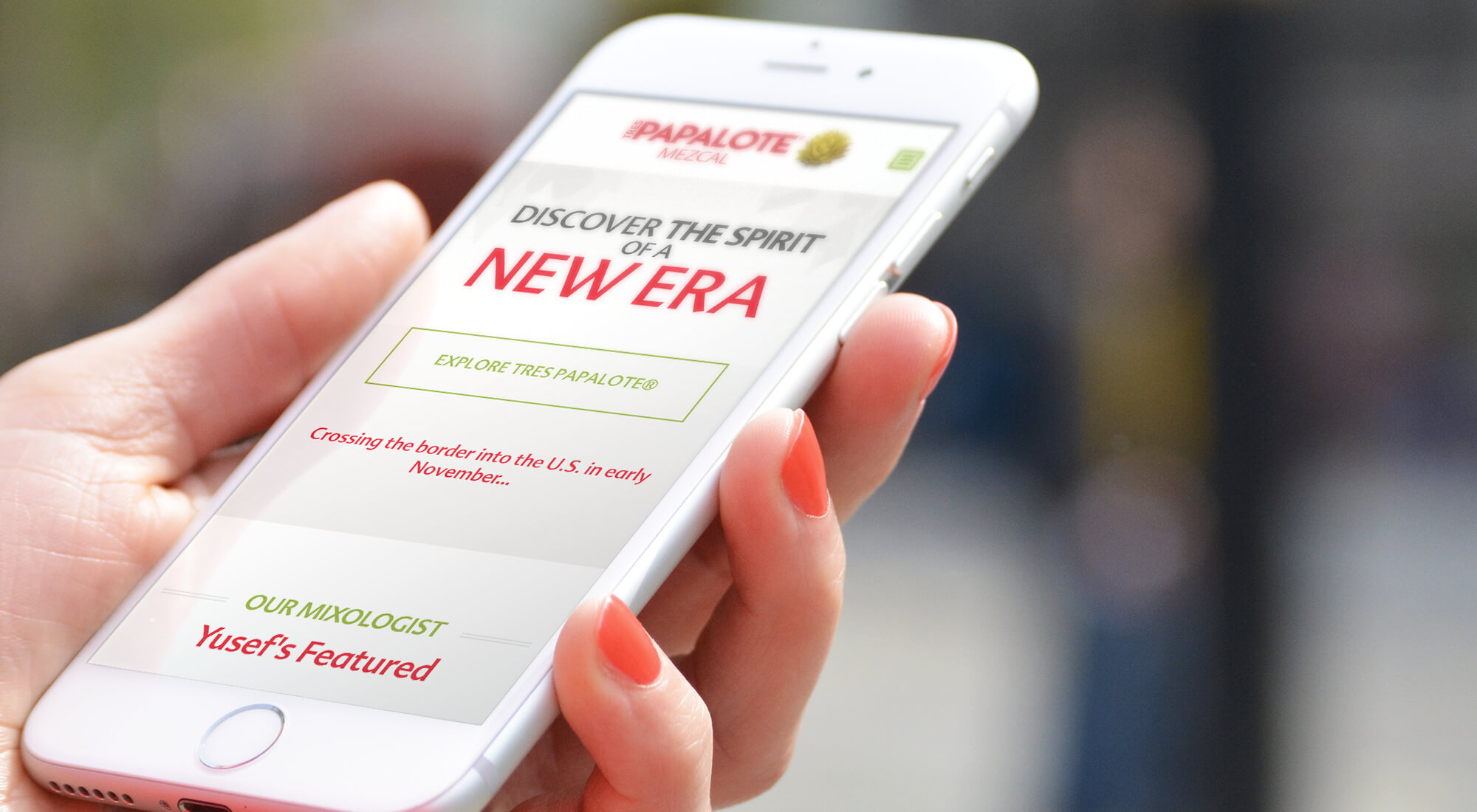 A person holds a smartphone displaying the Papalote Mezcal website, featuring promotional text about its U.S. launch and a section highlighting their mixologist.