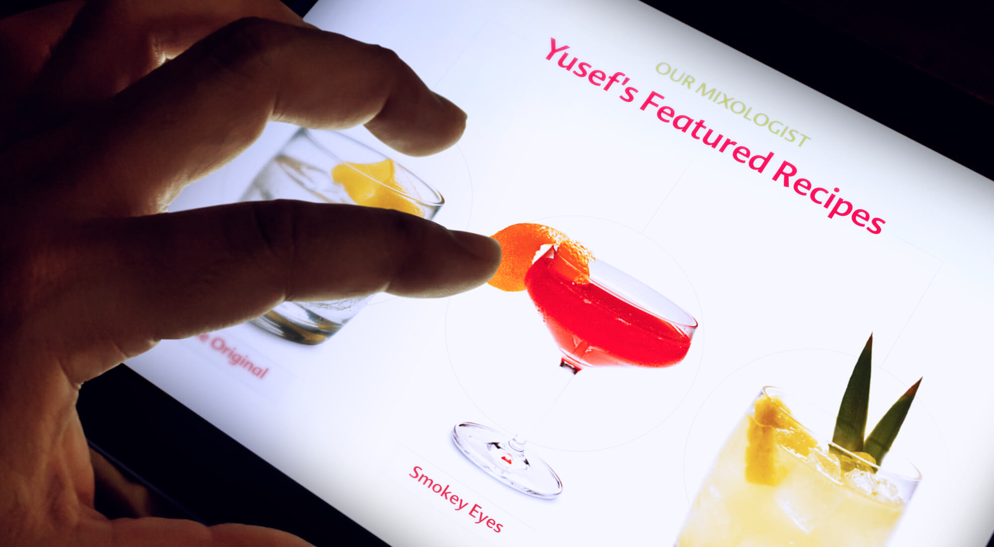 A hand interacts with a touchscreen displaying cocktail recipes, including drinks called "Smoky Eyes" and "Yusef's Original.
