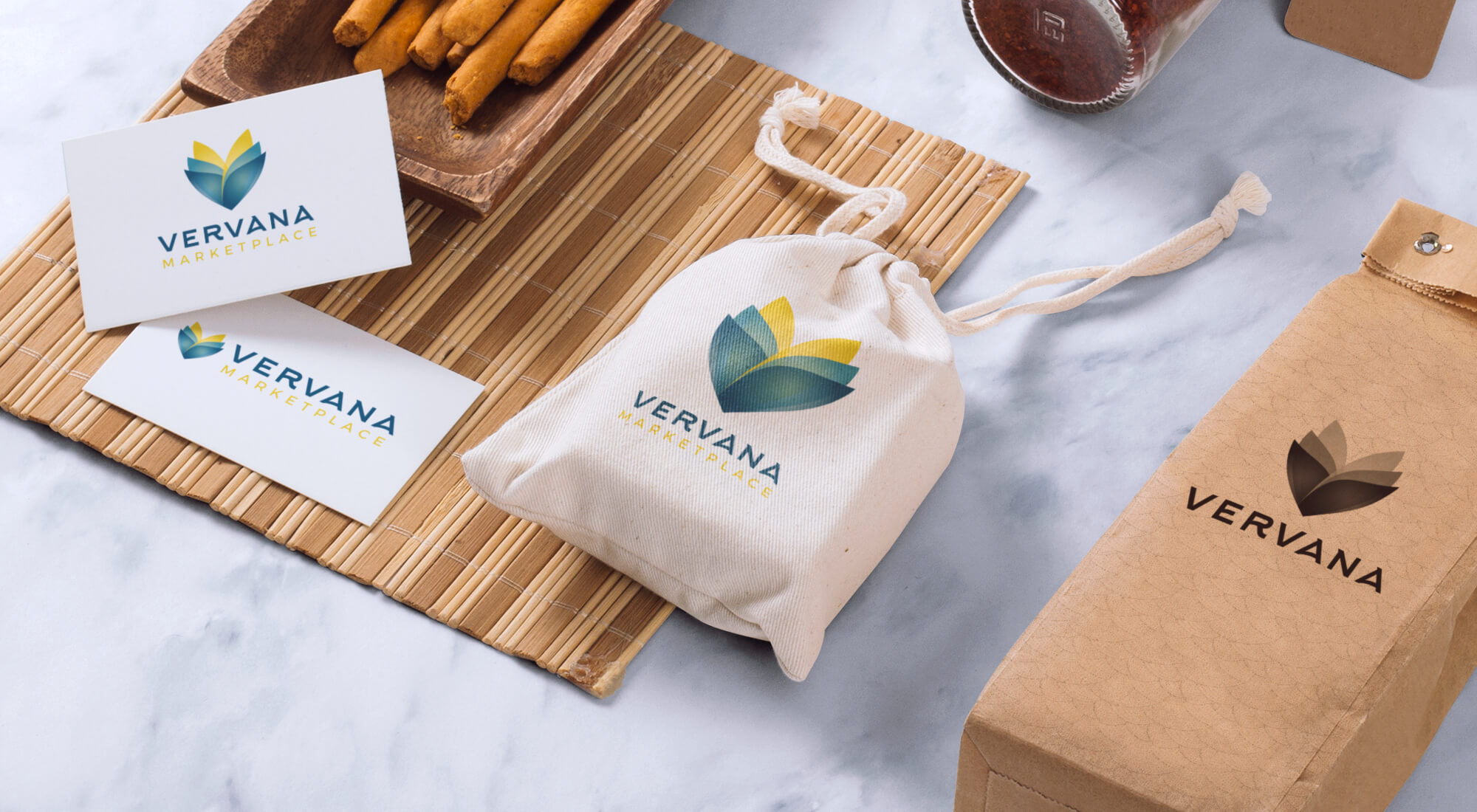 Branded items with the Vervana Marketplace logo, including business cards, a cloth pouch, and a cardboard box, are arranged on a marble surface with food and a bamboo placemat.