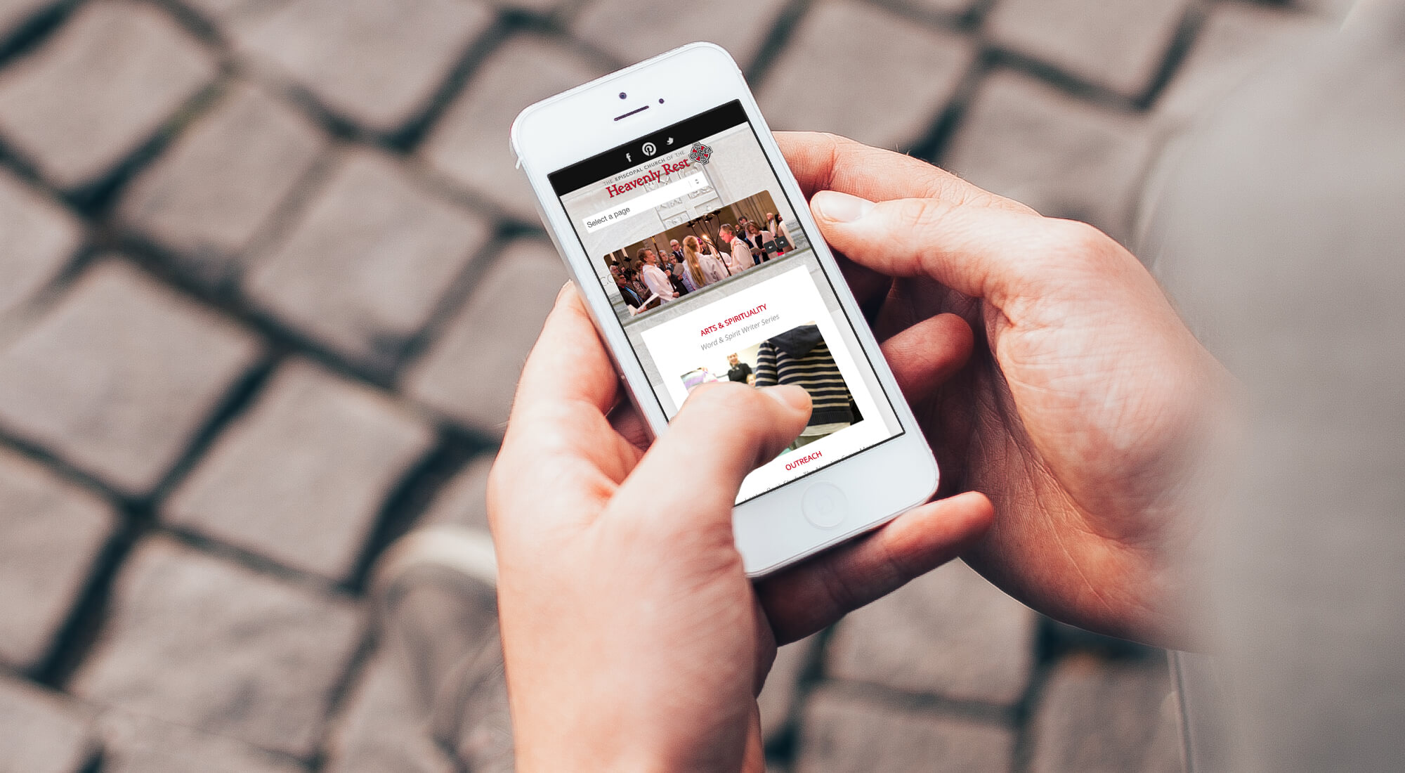 Person holds a white smartphone displaying a website while standing on a cobblestone surface, using their thumb to navigate the screen.