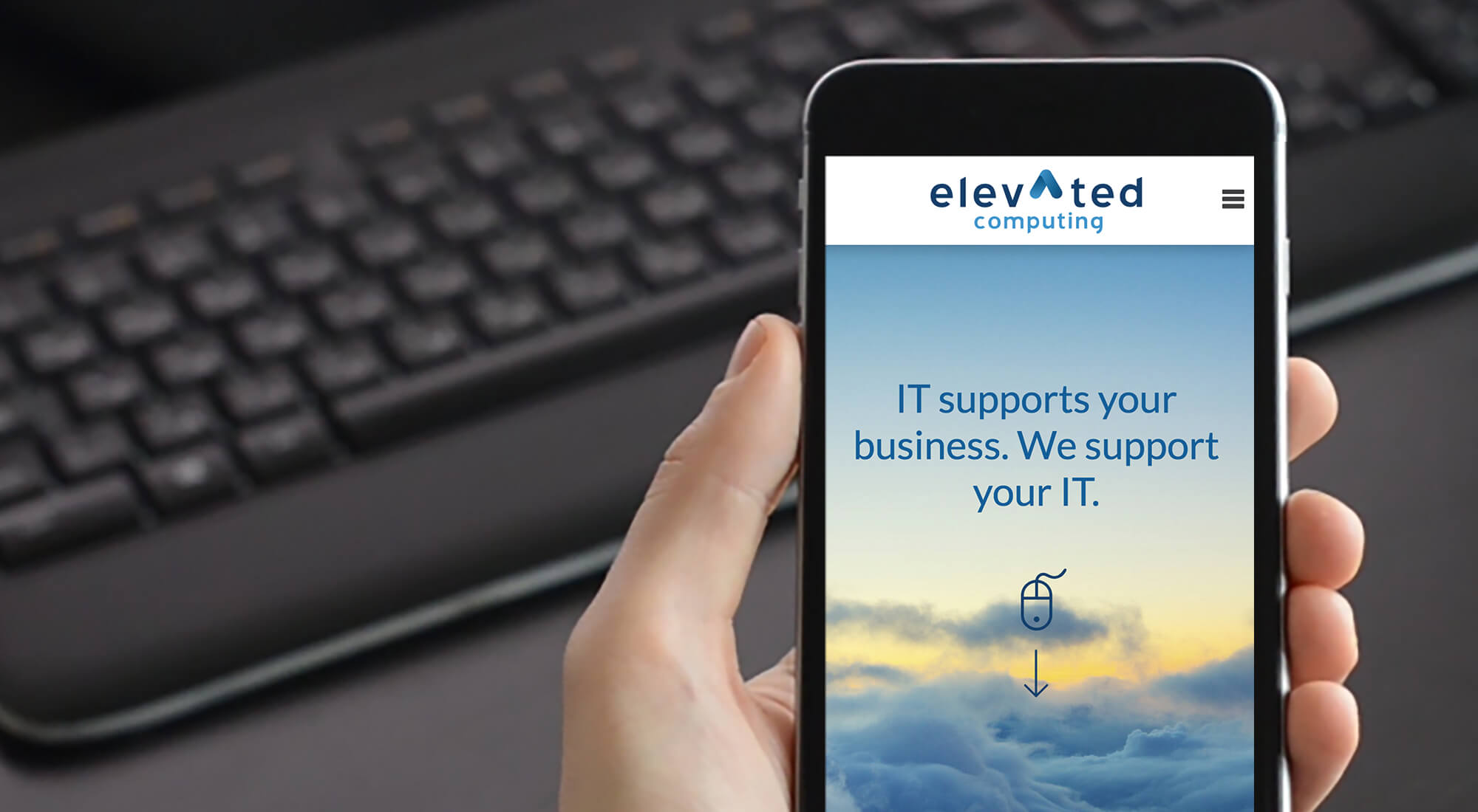 A hand holds a smartphone displaying the Elevated Computing website, with the message "IT supports your business. We support your IT." A keyboard is blurred in the background.