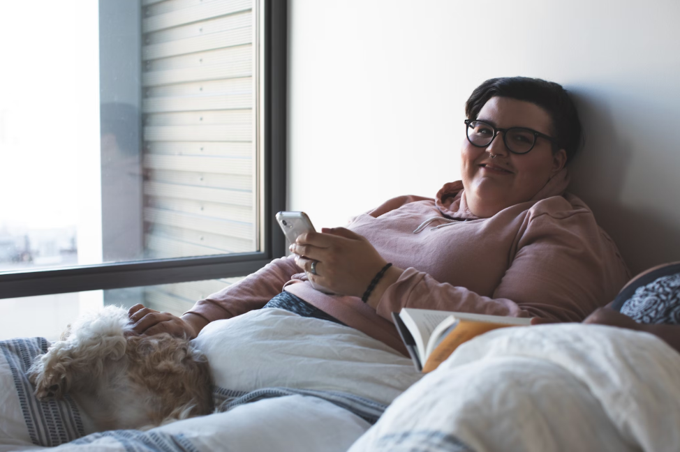 Person in a pink hoodie lying on a bed by a window, holding a phone, with their loyal dog resting nearby and an open book in the foreground—a cozy scene fit for any bebqueen.