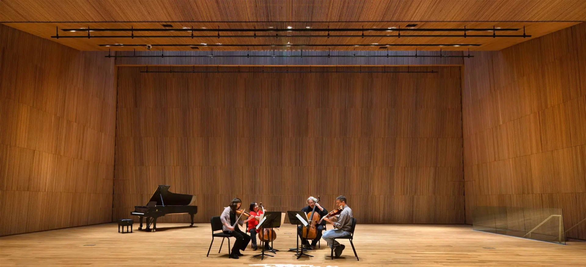 Four musicians sit in a circle playing string instruments on a wooden stage, with a grand piano in the background—a perfect scene for inspiring music website design. A large wooden wall rises behind them.