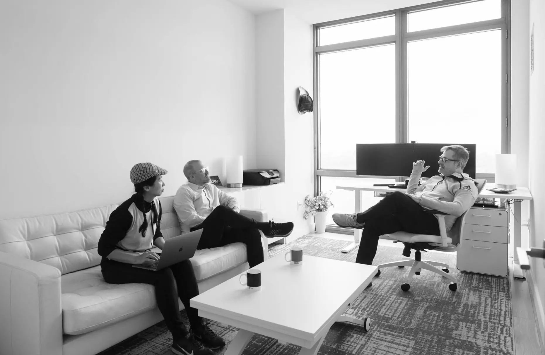 Three people are having a discussion in a modern office; two are seated on a couch with a laptop, and one is in an office chair near a desk with a computer monitor.