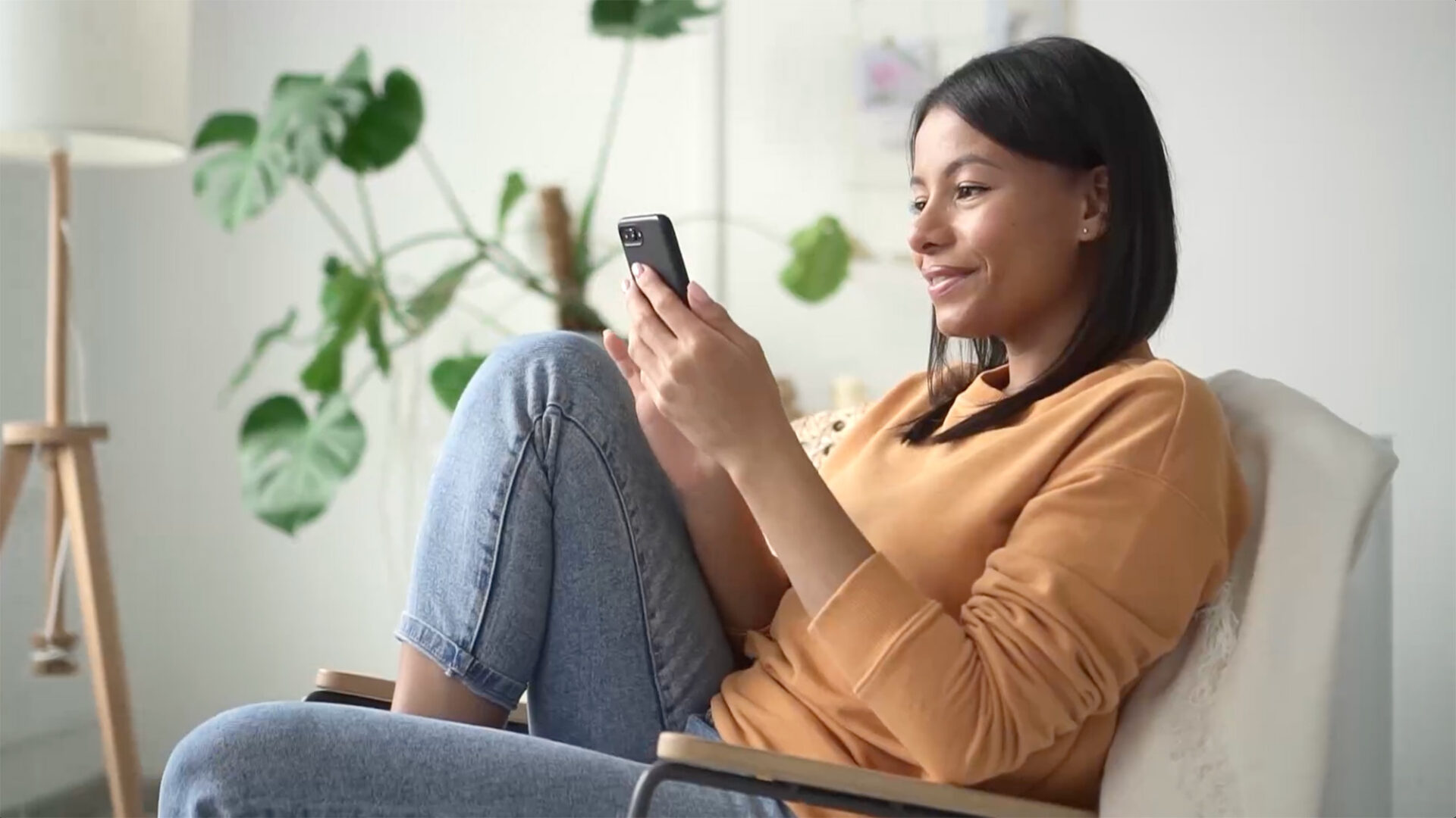 A woman in a tan sweater sits in a chair, smiling while looking at her smartphone—perhaps exploring a cancer care website design. A large leafy plant is in the background.