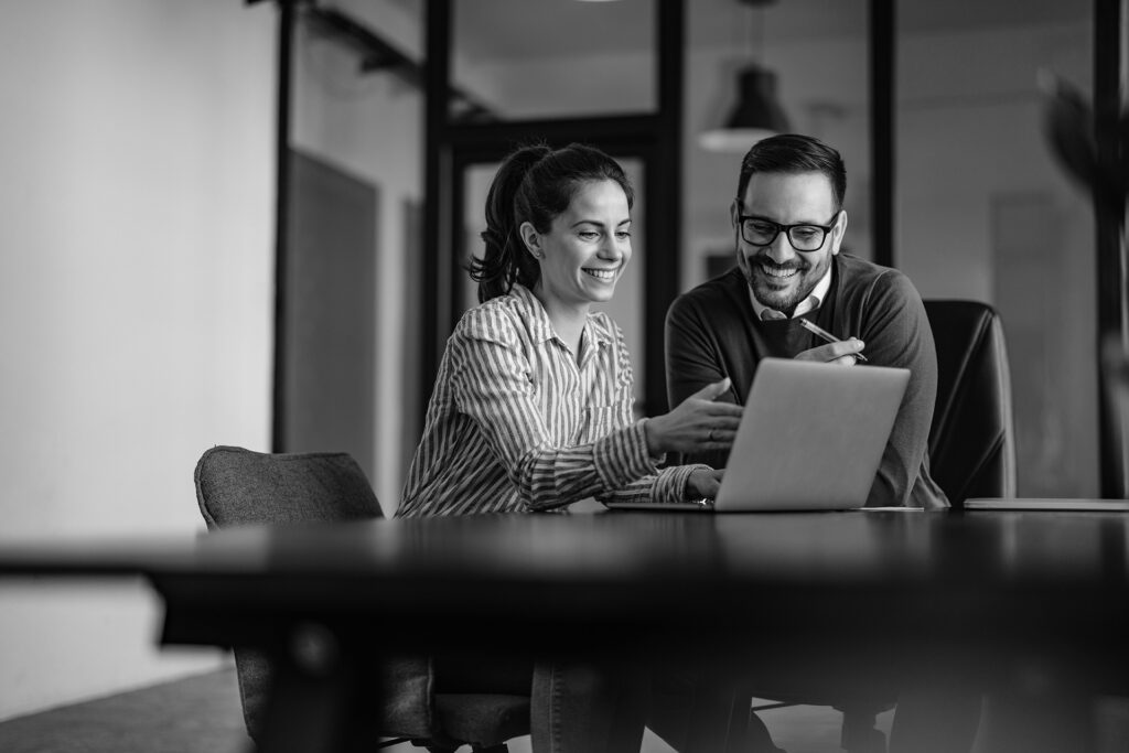 Two people sit at a table, smiling and looking at a laptop screen in an office setting, as they review the latest HR website design.