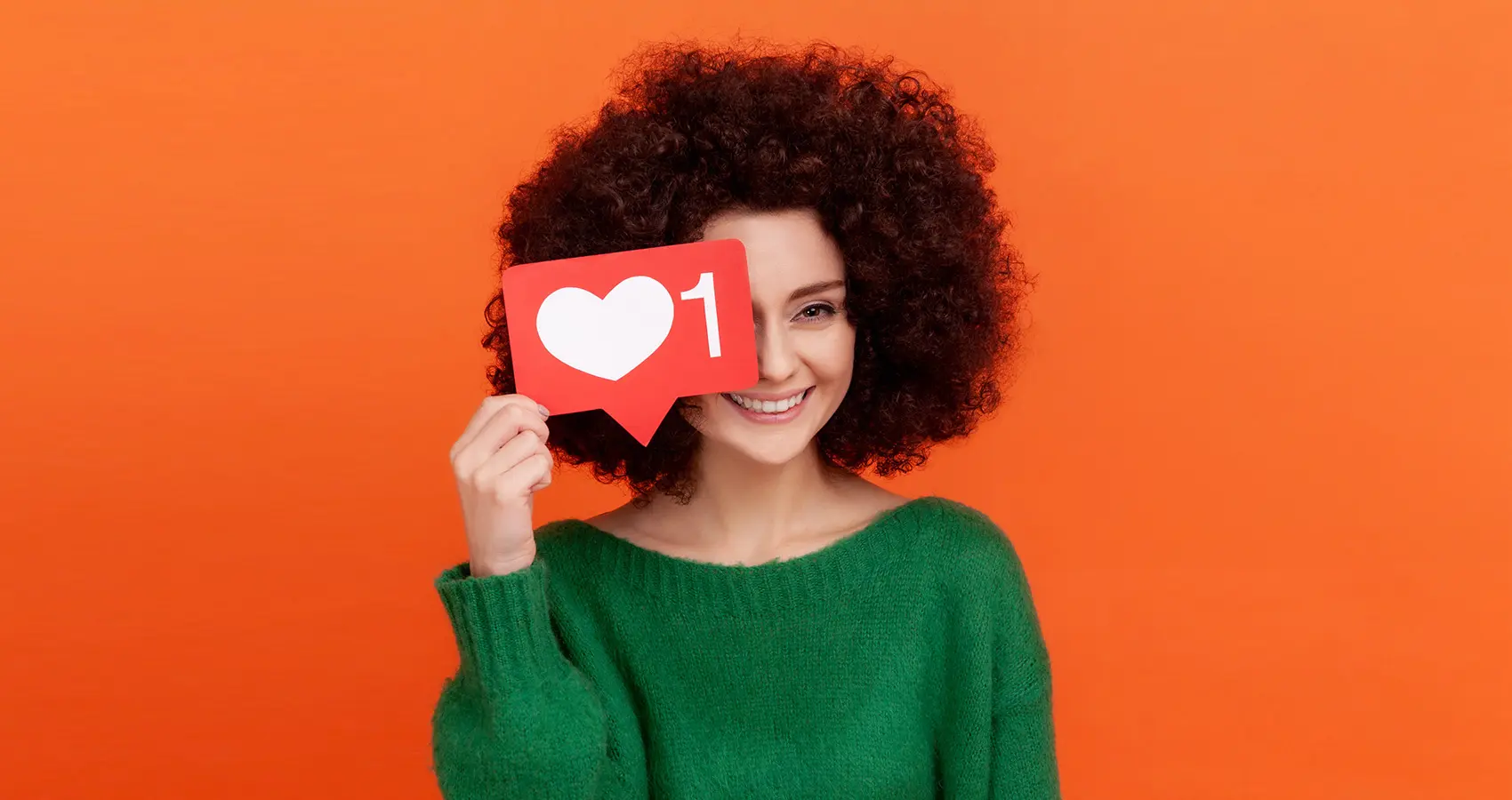 A woman with curly hair in a green sweater holds a red heart-shaped social media notification with the number 1, standing in front of an orange background.