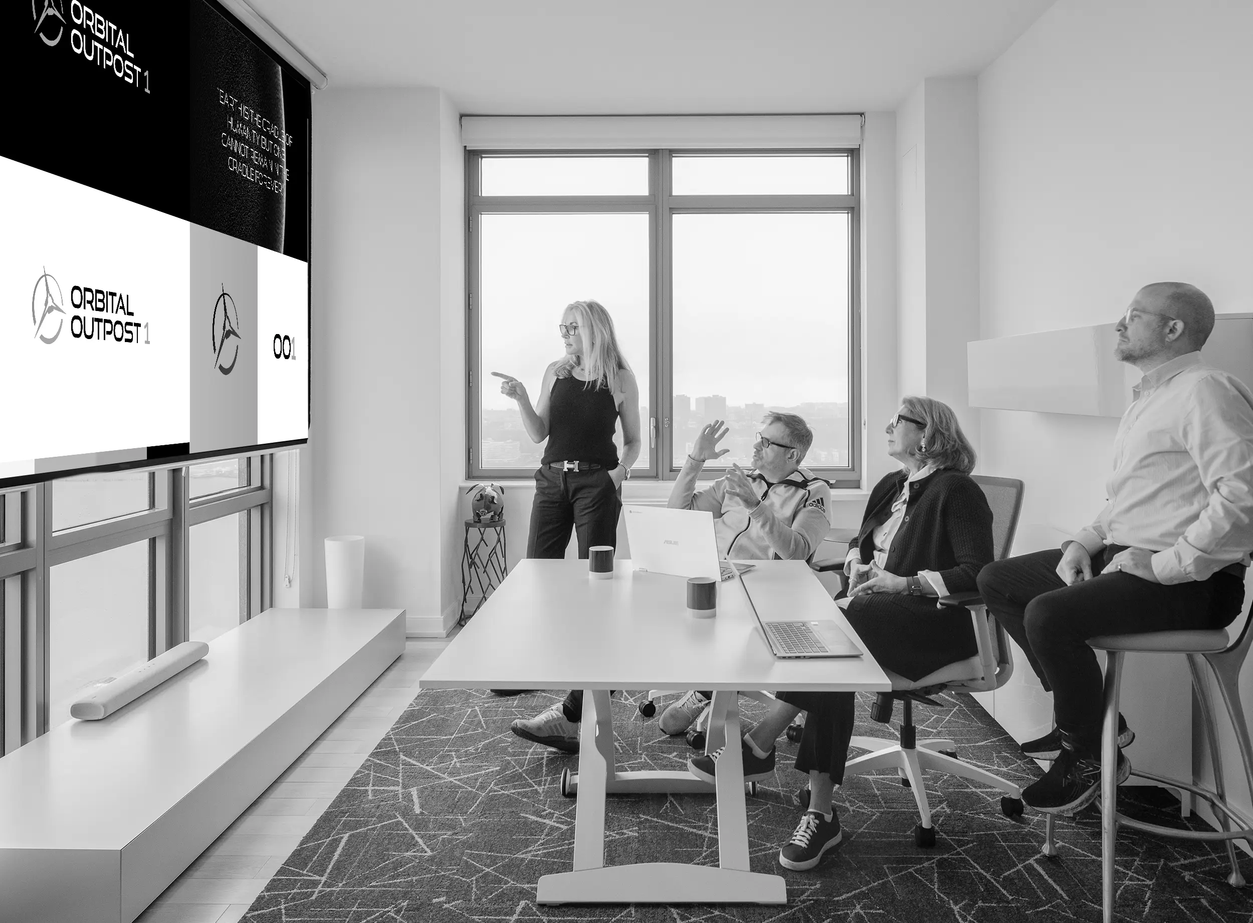 Four people in a modern office sit around a table, watching a woman present slides with logos on a large screen near a window.