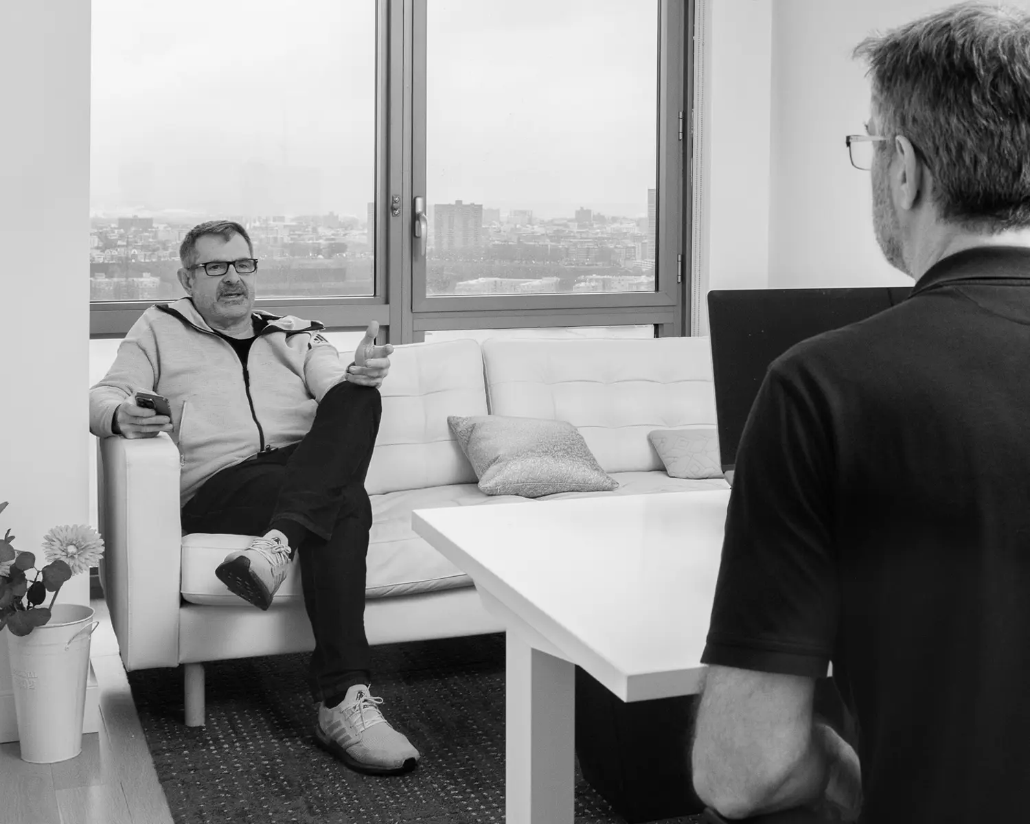 Two men are having a conversation in an office; one sits on a sofa holding a phone, while the other sits at a desk facing him.
