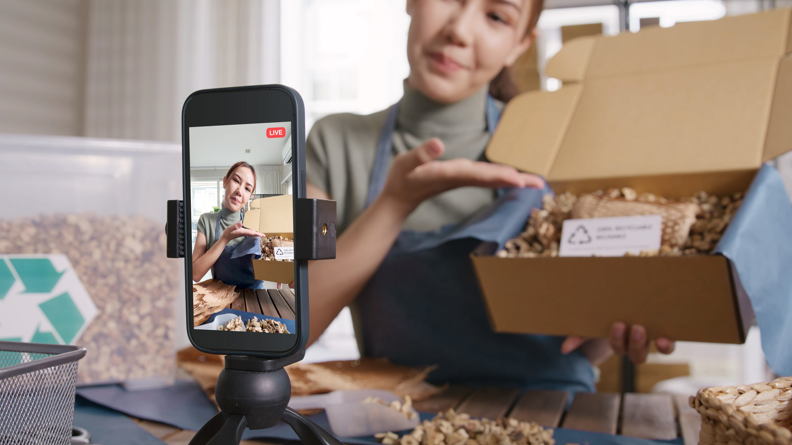 A person livestreams while holding an open cardboard box filled with packing material, demonstrating packaging to the camera on a smartphone tripod—showcasing Social Media Marketing For Small Business in action.