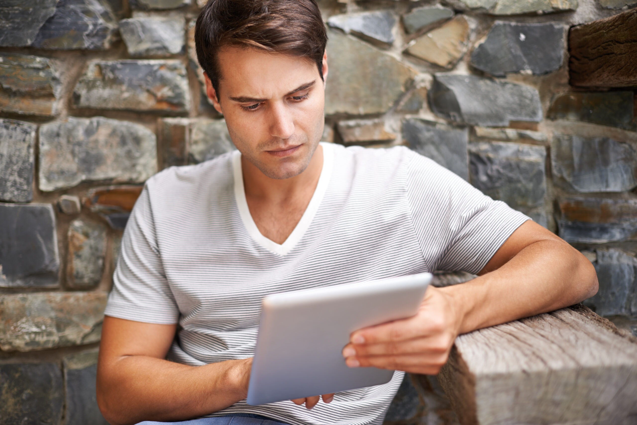 A man in a striped white t-shirt sits on a wooden bench outdoors, reading about Client Nightmares on his tablet with a stone wall in the background.