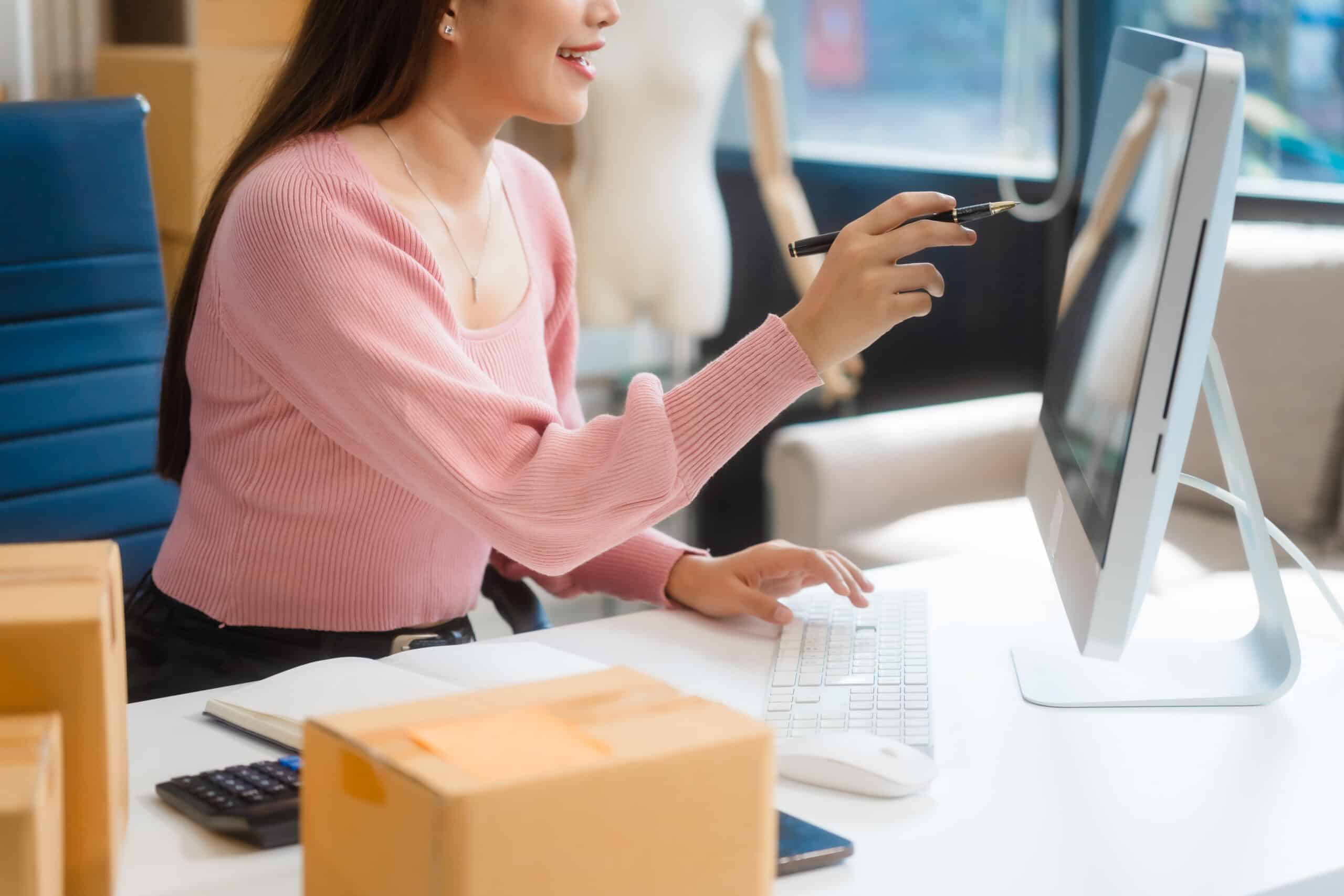 A woman in a pink sweater sits at a desk with a computer, pointing at the screen as she manages small business email marketing, surrounded by cardboard boxes and office supplies.