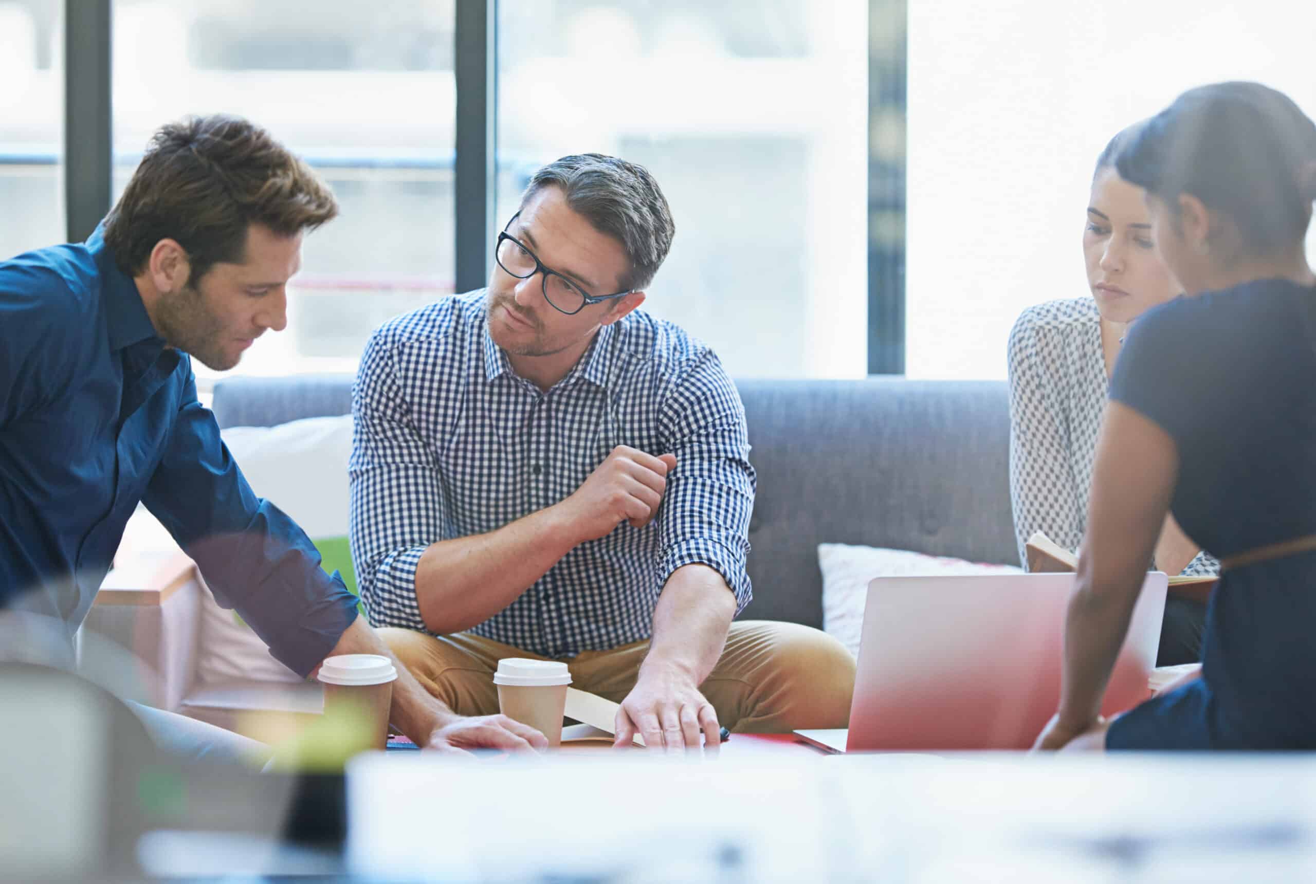 Four people sit together in an office setting, discussing documents and laptops over coffee as they review important Questions To Ask Your Web Agency.