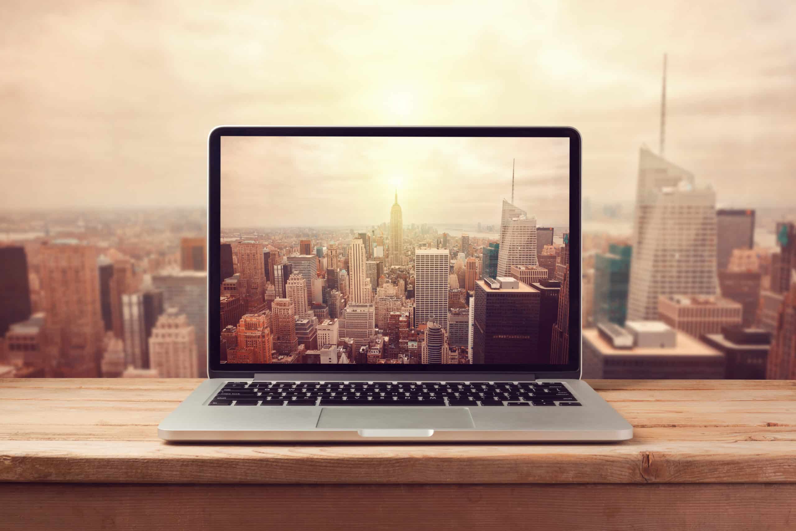 A laptop on a wooden table displays a city skyline on its screen, with a similar city view seen in the background, suggesting a blend of virtual and real perspectives.