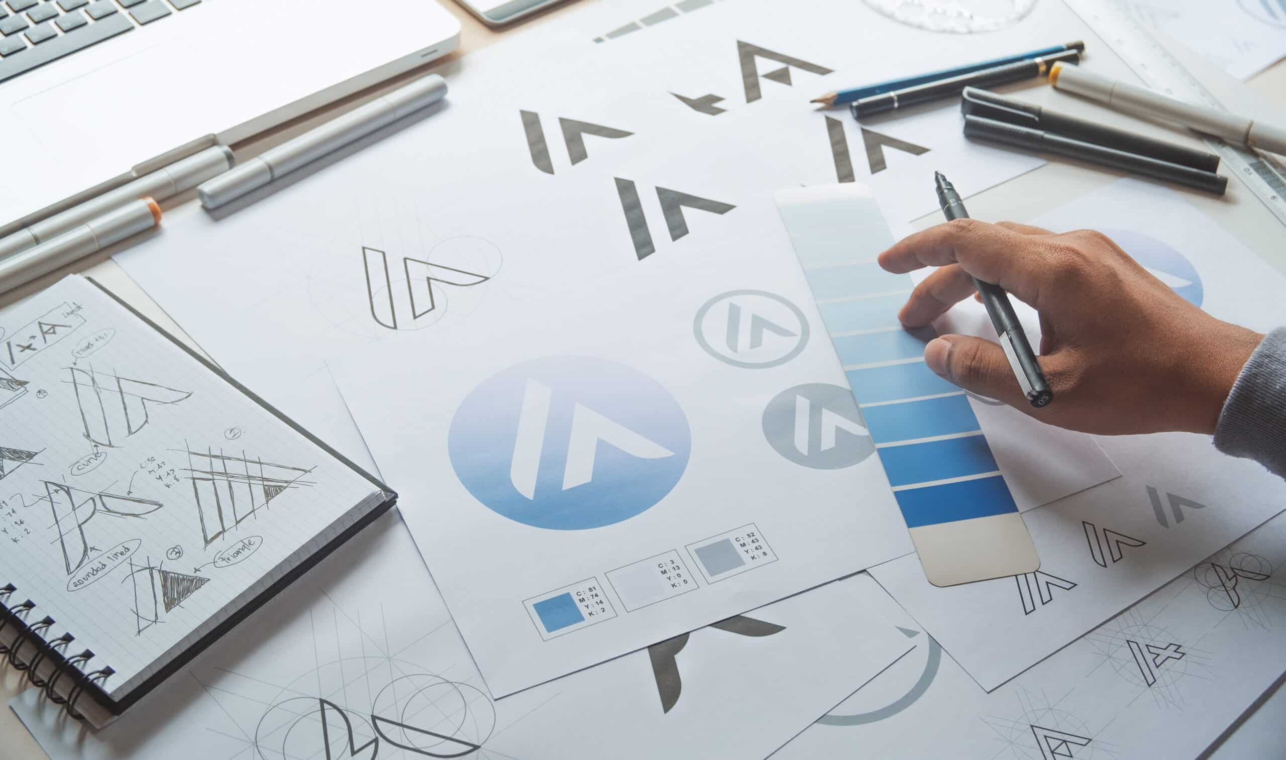 Designer reviewing blue color swatches and logo sketches on paper, with pens, a notebook, and a laptop on the workspace.