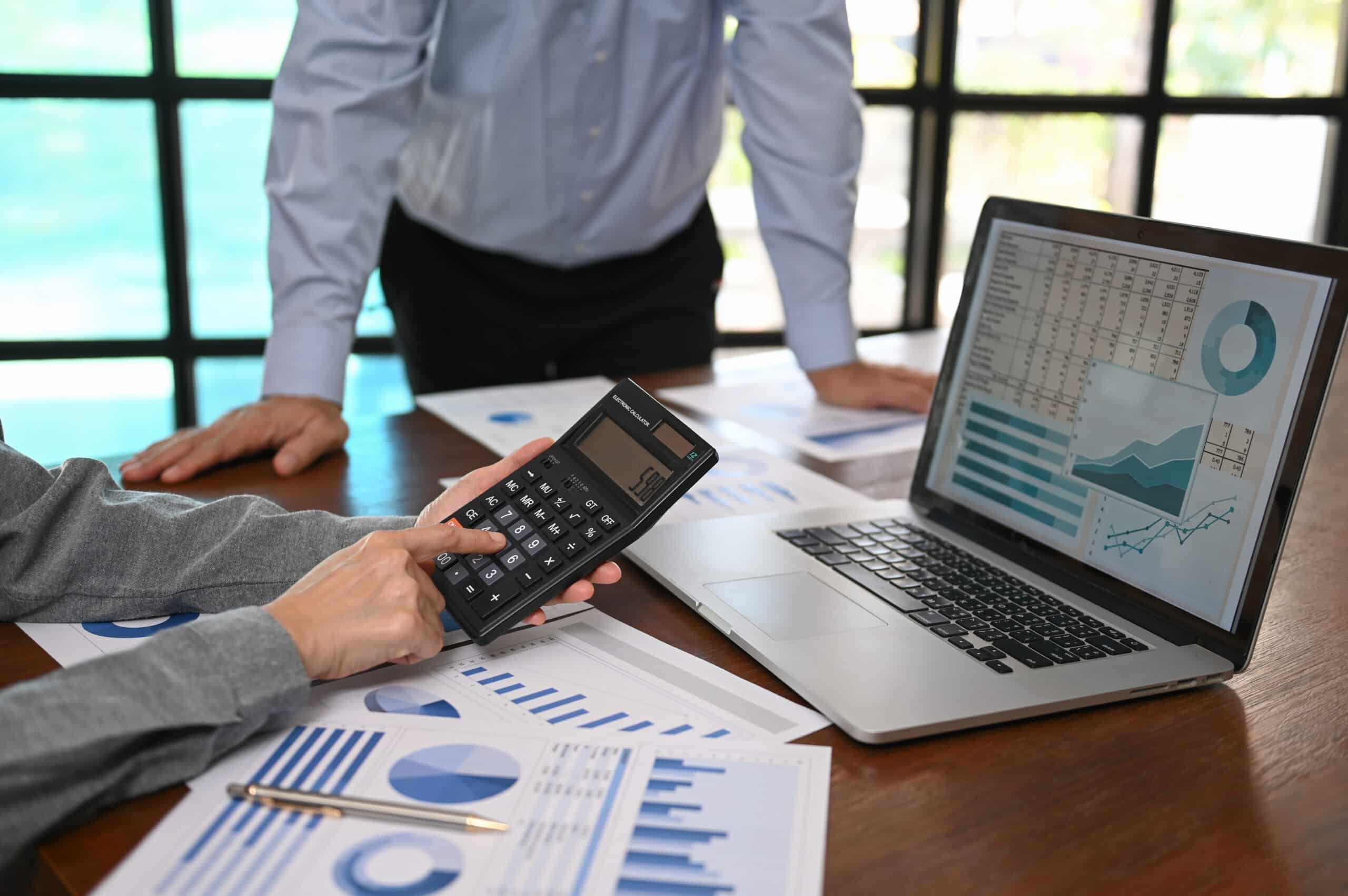 Two people reviewing financial charts and graphs on paper and a laptop, with one person using a calculator at a wooden desk—analyzing data critical for SEO For Accountants success.