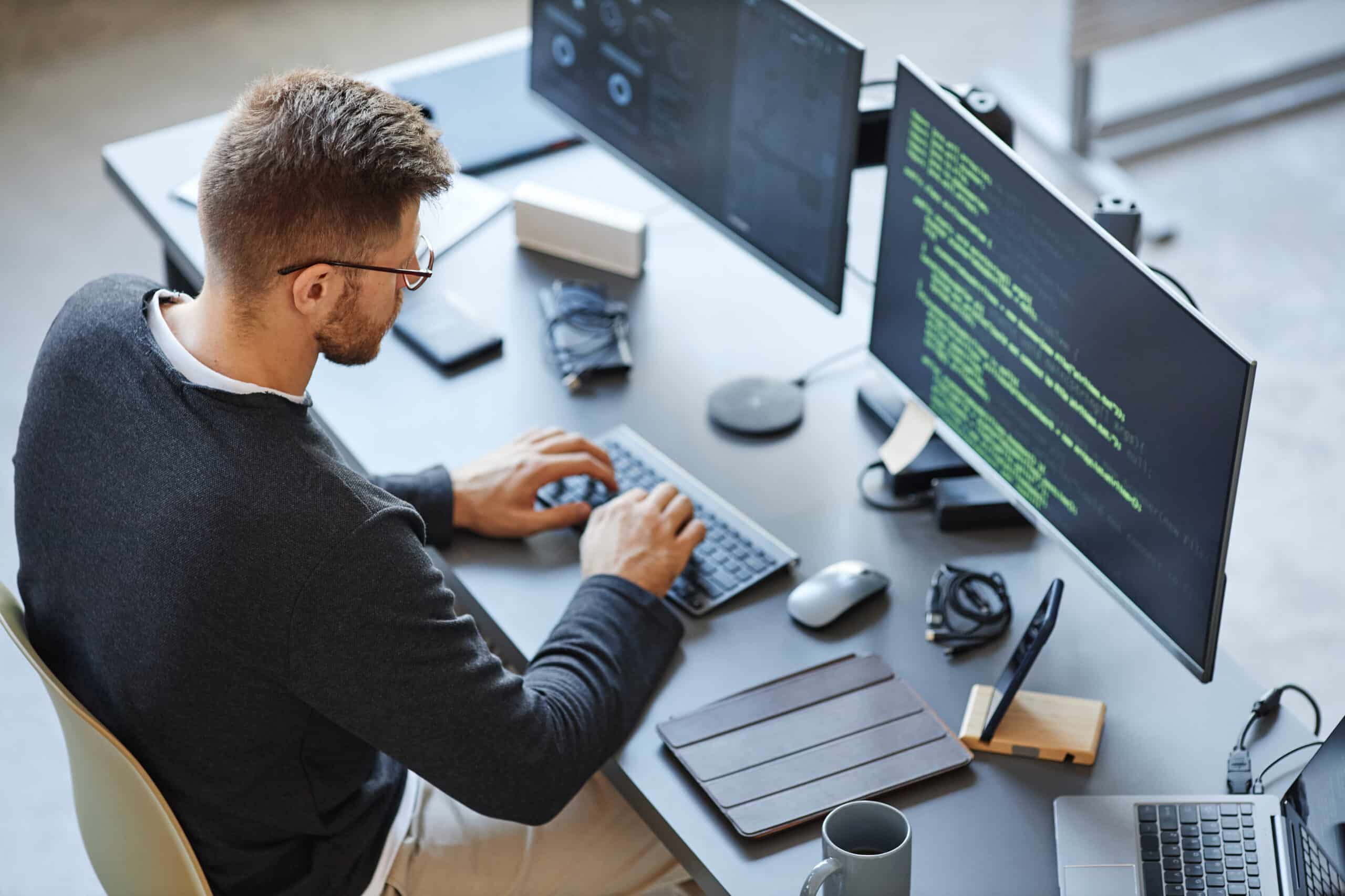 Man working at a desk with two monitors displaying code, typing on a keyboard. Various office items and electronics are on the desk.
