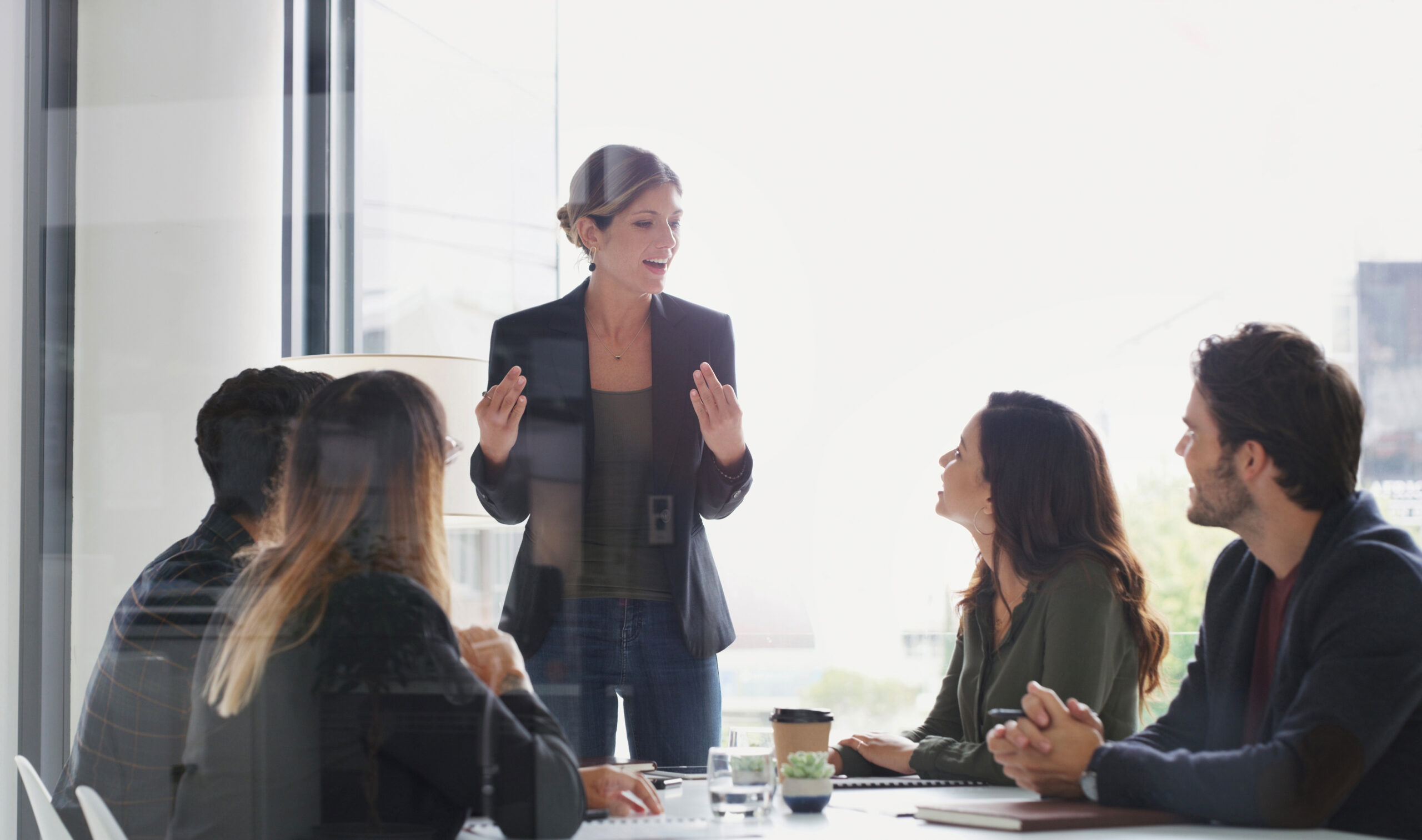 A woman stands and speaks to four seated colleagues around a conference table in a modern office setting with large windows.